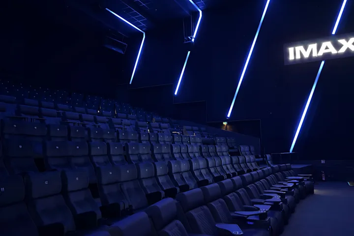 Moderner IMAX-Kinosaal mit Reihen aus dunklen, gepolsterten Sitzen, blauer LED-Lichtinszenierung an Wänden und Decke sowie leuchtendem IMAX-Logo im Hintergrund.