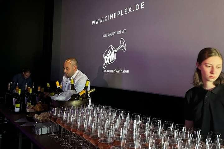 „Kinoleinwand zeigt die Begrüßung zu Kino et Vino, davor stehen Gäste mit Weingläsern“