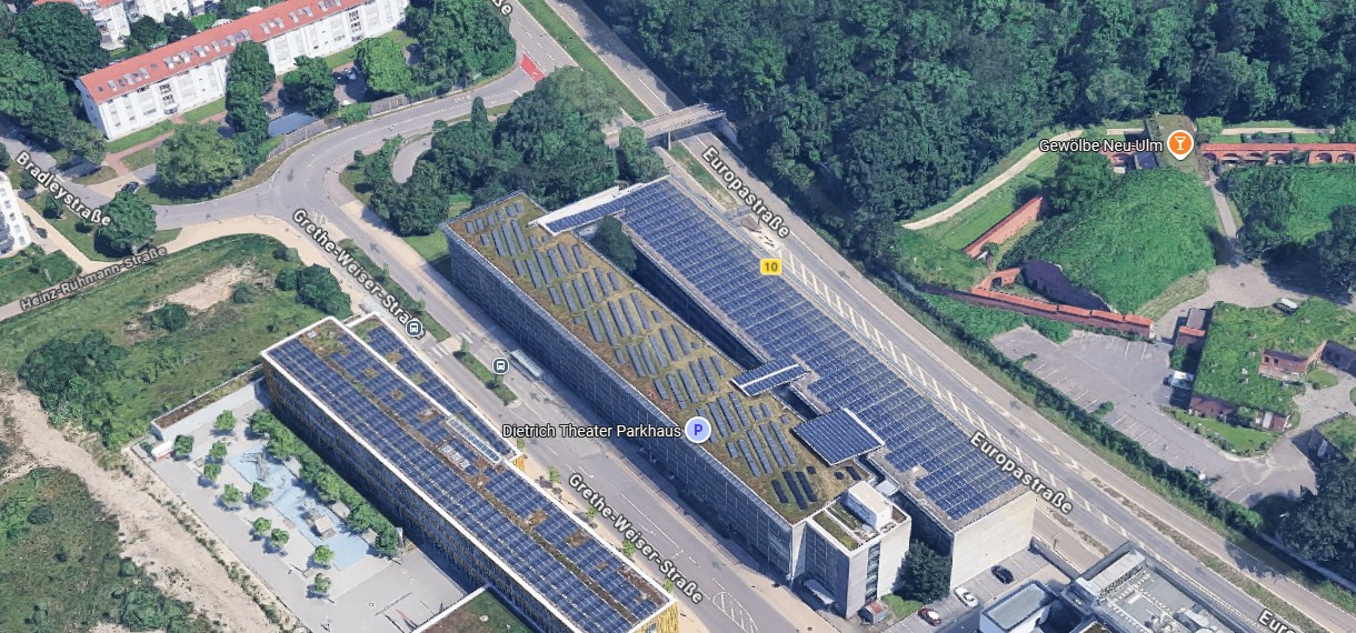 Schräg-Luftaufnahme eines langen Parkhauses mit begrüntem und mit Solarpanels bedecktem Dach. Das Gebäude liegt an der Europastraße in Neu-Ulm, umgeben von Wohnhäusern, Bäumen und alten Festungsanlagen.