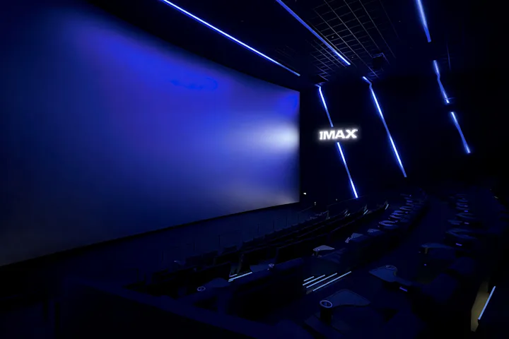 Dunkler IMAX-Kinosaal mit großer, leicht gebogener Leinwand, blauer LED-Beleuchtung an Wänden und Decke sowie sichtbaren Sitzreihen im Vordergrund.
