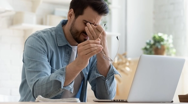 Мужчина сидит за столом перед ноутбуком Мужчина перед ноутбуком с признаками дискомфорта в глазах