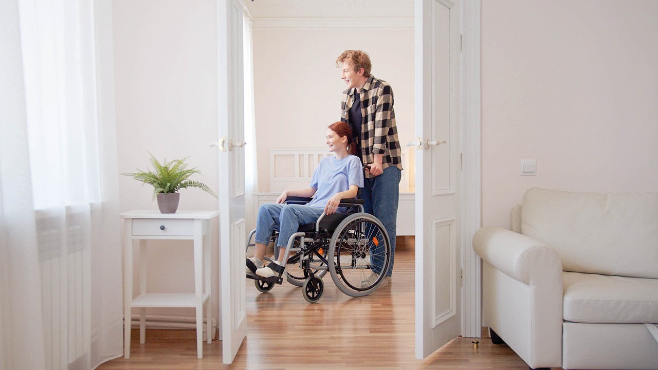 Junge Frau im Rollstuhl wird in der Wohnung unterstützt.