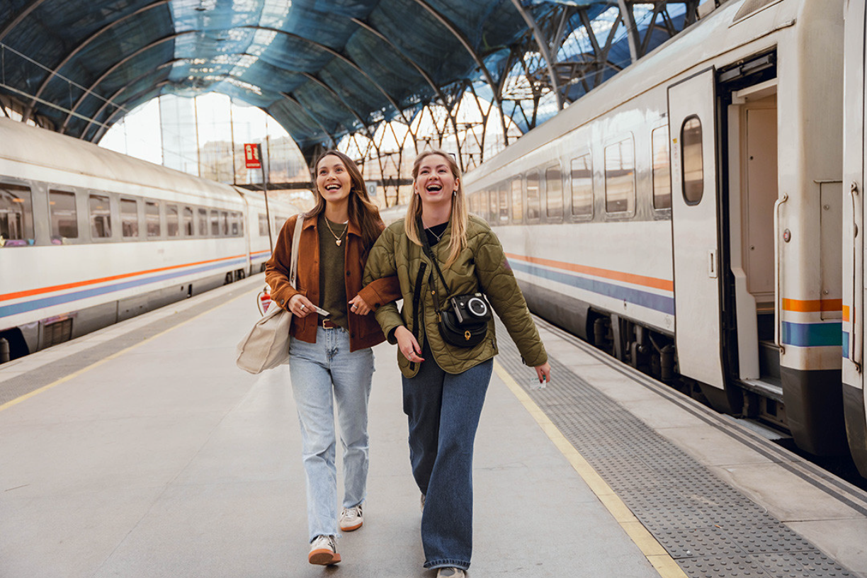 Zwei Frauen halten sich lächelnd an den Armen und spazieren am Bahnsteig