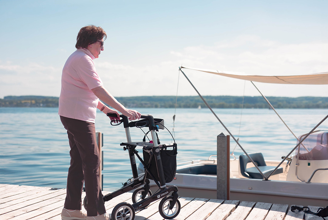 Eine Seniorin steht mit einem Rollator auf einem Steg am Wasser