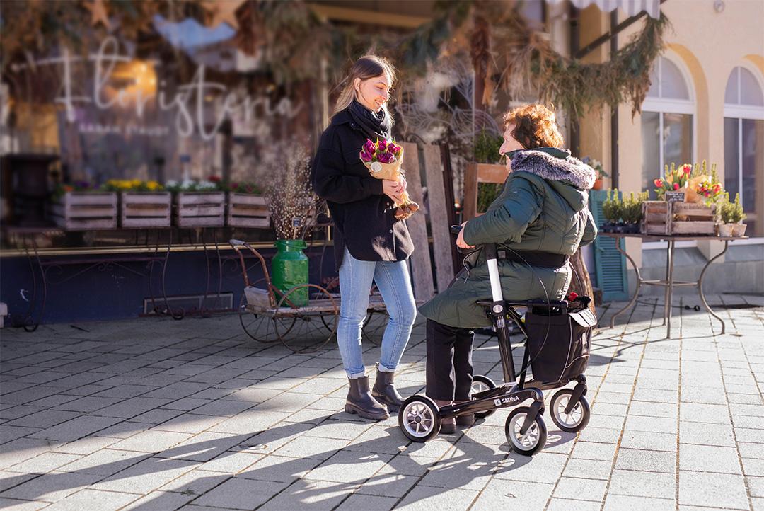 Eine Seniorin sitzt auf einem Rollator vor einem Blumenladen und eine junge Frau hält einen Blumenstrauß.