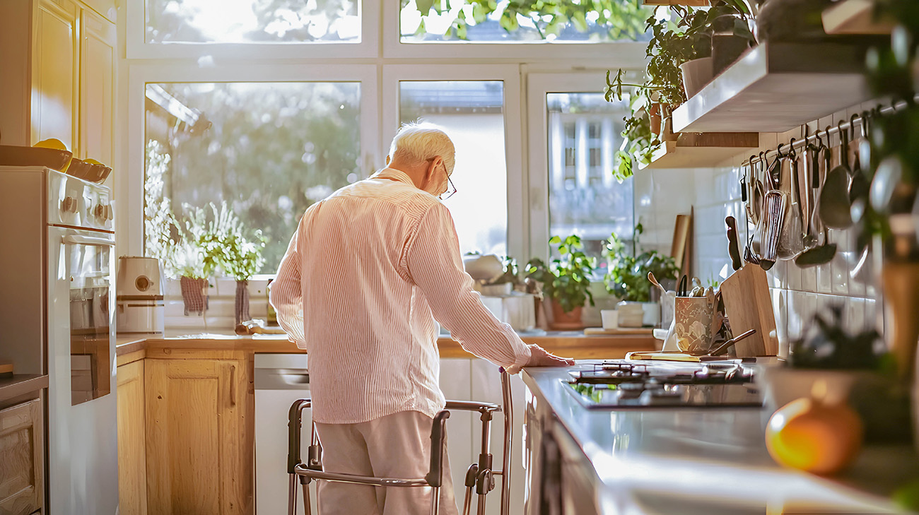 Ein Senior mit einem Gehgestell steht in der Küche und hält sich an der Arbeitsfläche