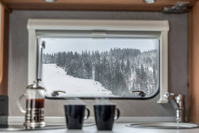 Tassen und Kaffee vor einem Wohnmobilfenster mit Blick auf Winterlandschaft