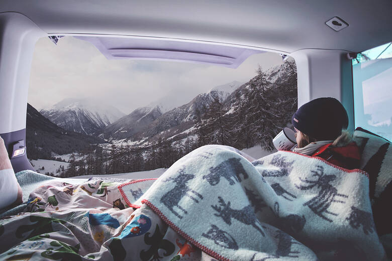 Frau sitzt mit Decke und einer Tasse Kaffee im Wohnmobil und schaut auf eine Winterlandschaft