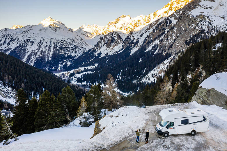 Familie steht vor dem Wohnmobil und schaut auf eine verschneite Alpenlandschaft