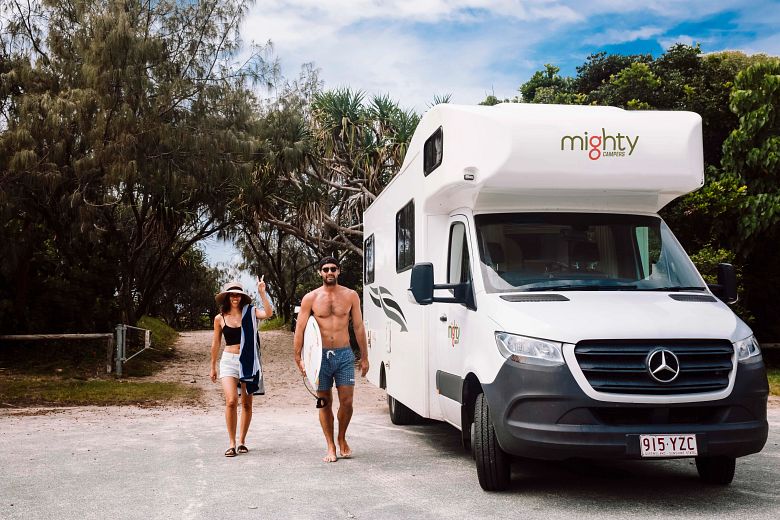 Paar mit Surfboard vor großem Mighty Wohnmobil