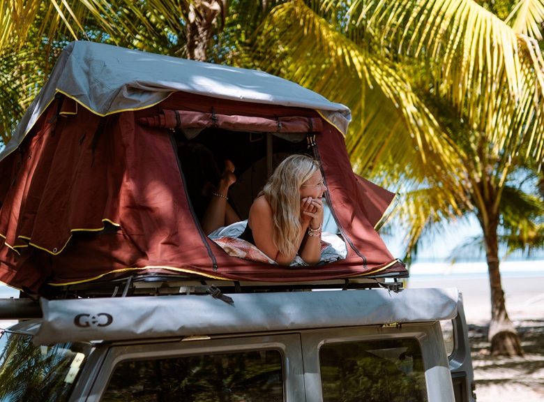 Frau liegt in einem Dachzelt auf einem Camper unter Palmen