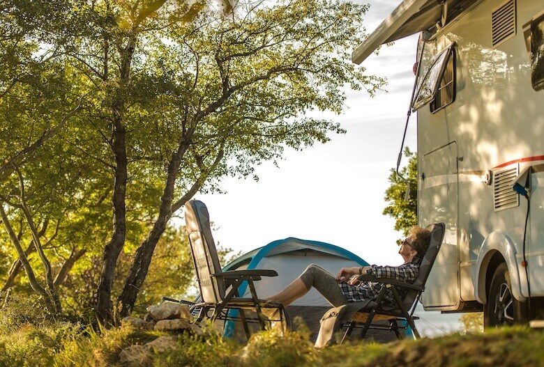 Camping for seniors: an older lady relaxes on camping chairs in front of her RV.