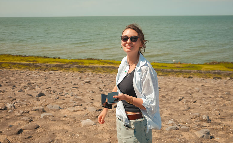Frau steht am Meer, mit zwei Smartphones in der Hand, und lächelt in die Kamera