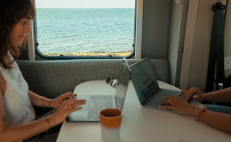 Frau arbeitet am Laptop und schaut aus dem Camper-Fenster auf das Meer