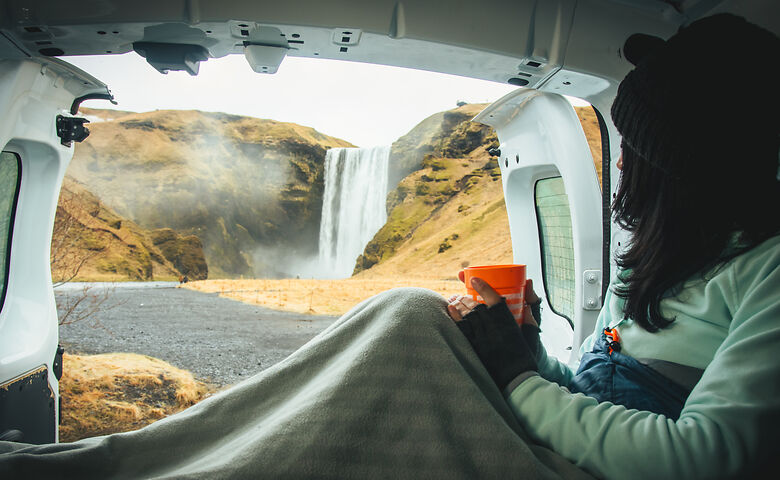 Een vrouw drinkt koffie in de camper en kijkt naar de Skógafoss in IJsland