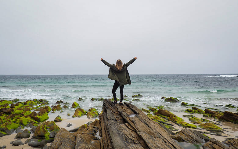 Frau steht an einem Strand in irland und hebt die Arme in die Luft