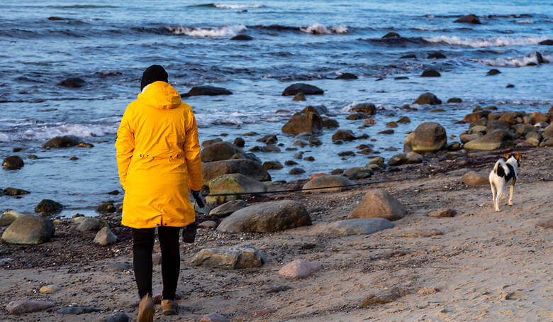 Frau mit gelber Regenjacke geht am Meer mit Hund spazieren