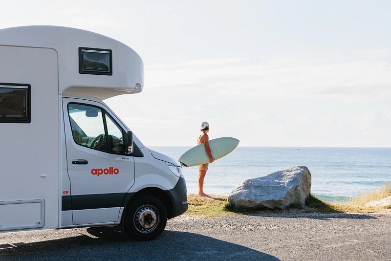 Mann mit Surfbrett neben Apollo-Wohnmobil am Meer