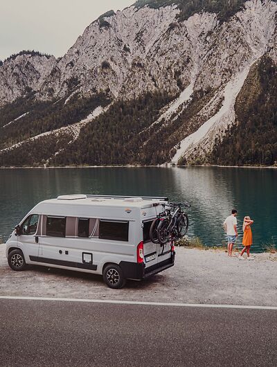 Paar steht mit einem Campervan an einem Bergsee