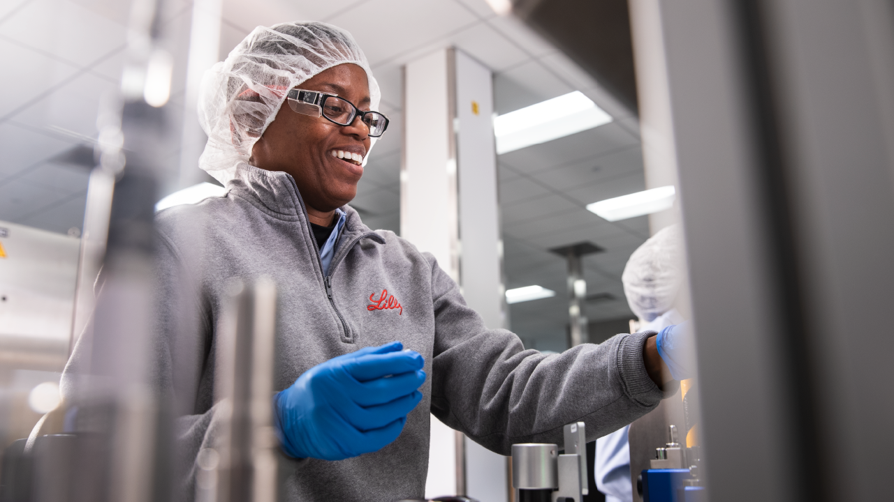 Black woman wearing Lilly pullover, hair net, and blue gloves works in lab