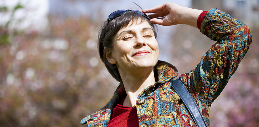 Middle aged woman stands outdoors with sunglasses on top of her head and smiles with eyes closed facing the sun