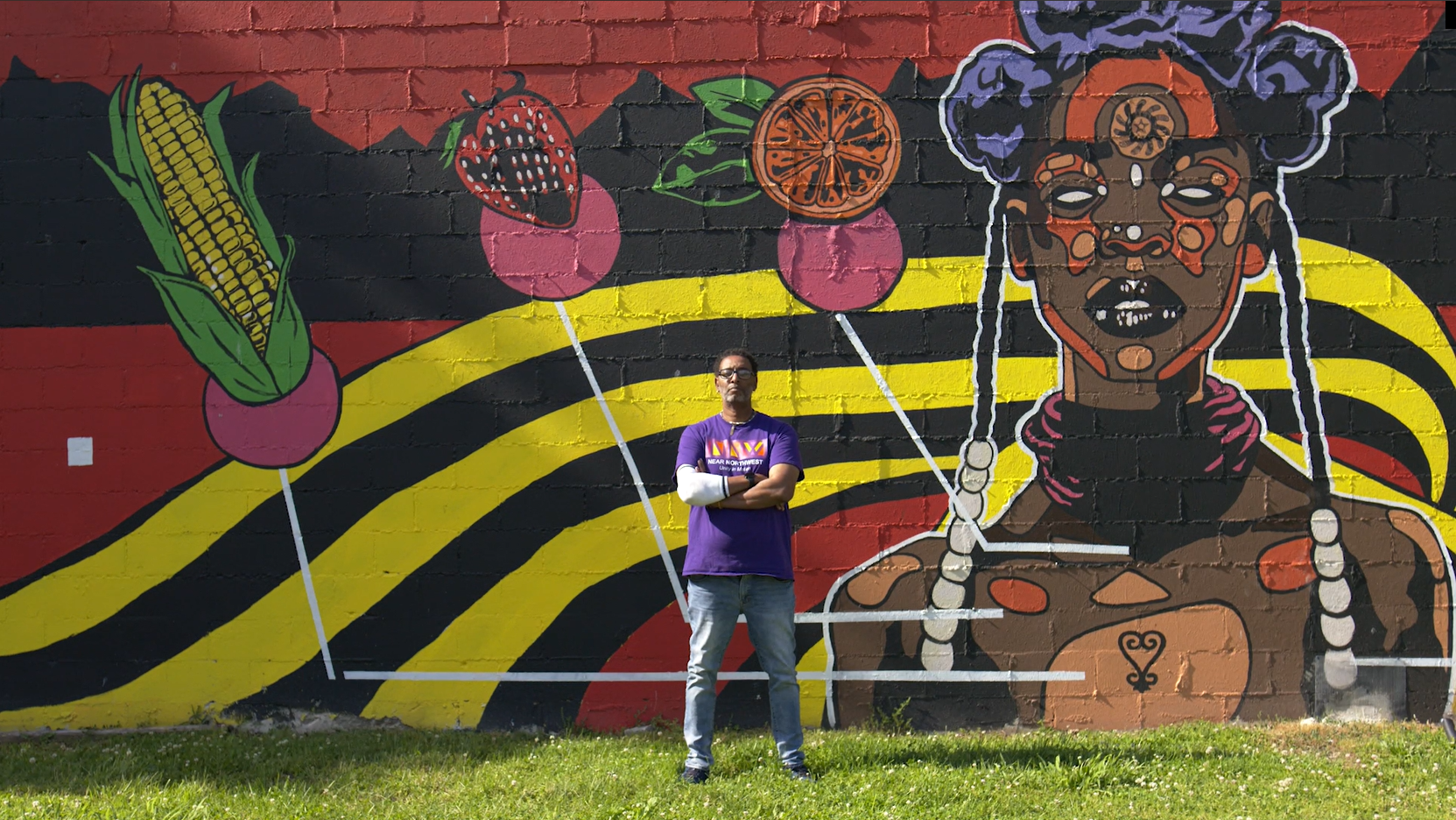 man standing outside with a vibrant african mural wall with a ear of corn, a strawberry and an orange slice. 