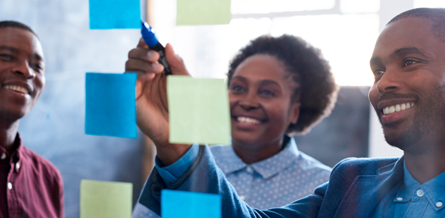 two Black men and one Black woman pointing to sticky notes on clear board