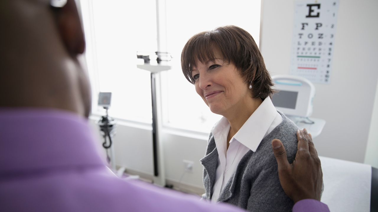 middle-aged white woman in doctor's office smiling, Black doctor pats patient's shoulder