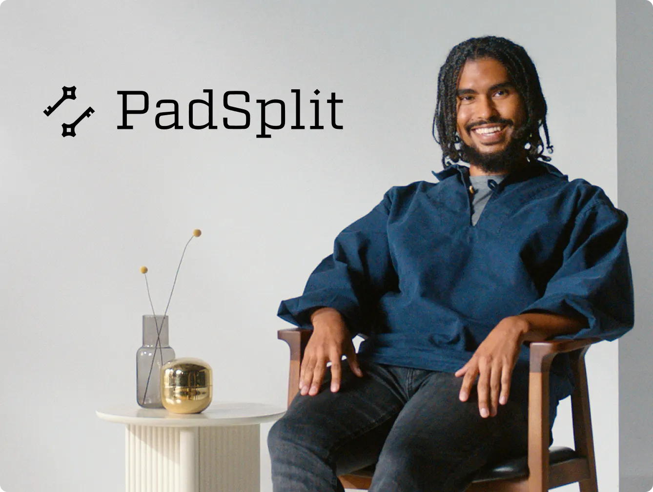 PadSplit advertisement featuring a smiling man with curly hair and a beard, wearing a dark blue shirt, sitting on a chair. Next to him is a small table with a vase and a decorative item. The PadSplit logo is prominently displayed at the top left of the image on a light gray background.