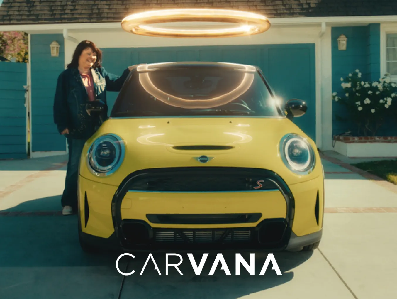 Carvana advertisement featuring a woman standing next to a yellow Mini Cooper parked in front of a blue house. A glowing halo is above the car. The Carvana logo is displayed at the bottom of the image.

