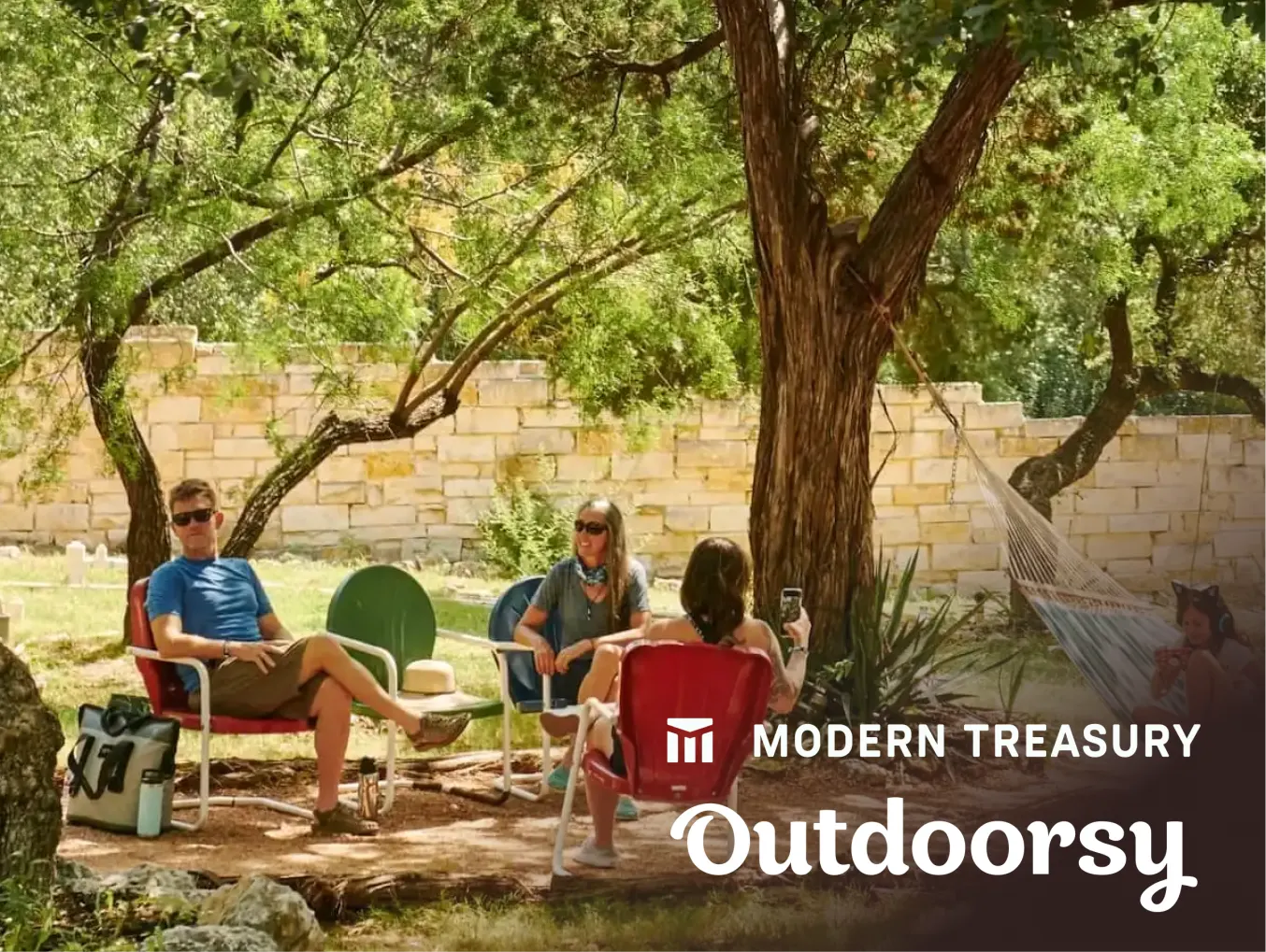 A group of people sitting outdoors in a shaded area with trees and a stone wall. They are relaxing on chairs and a hammock. The Modern Treasury and Outdoorsy logos are prominently displayed at the bottom right of the image.
