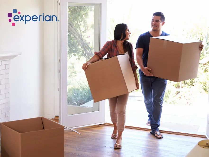 Smiling couple carrying moving boxes into a bright home, with the Experian logo in the corner.