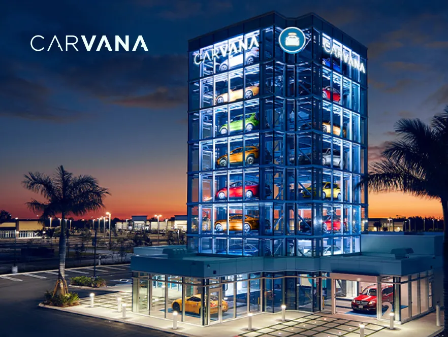 Carvana's illuminated multi-story glass car vending machine at sunset, showcasing various brightly colored vehicles stacked on display, with palm trees and a parking lot in the foreground.