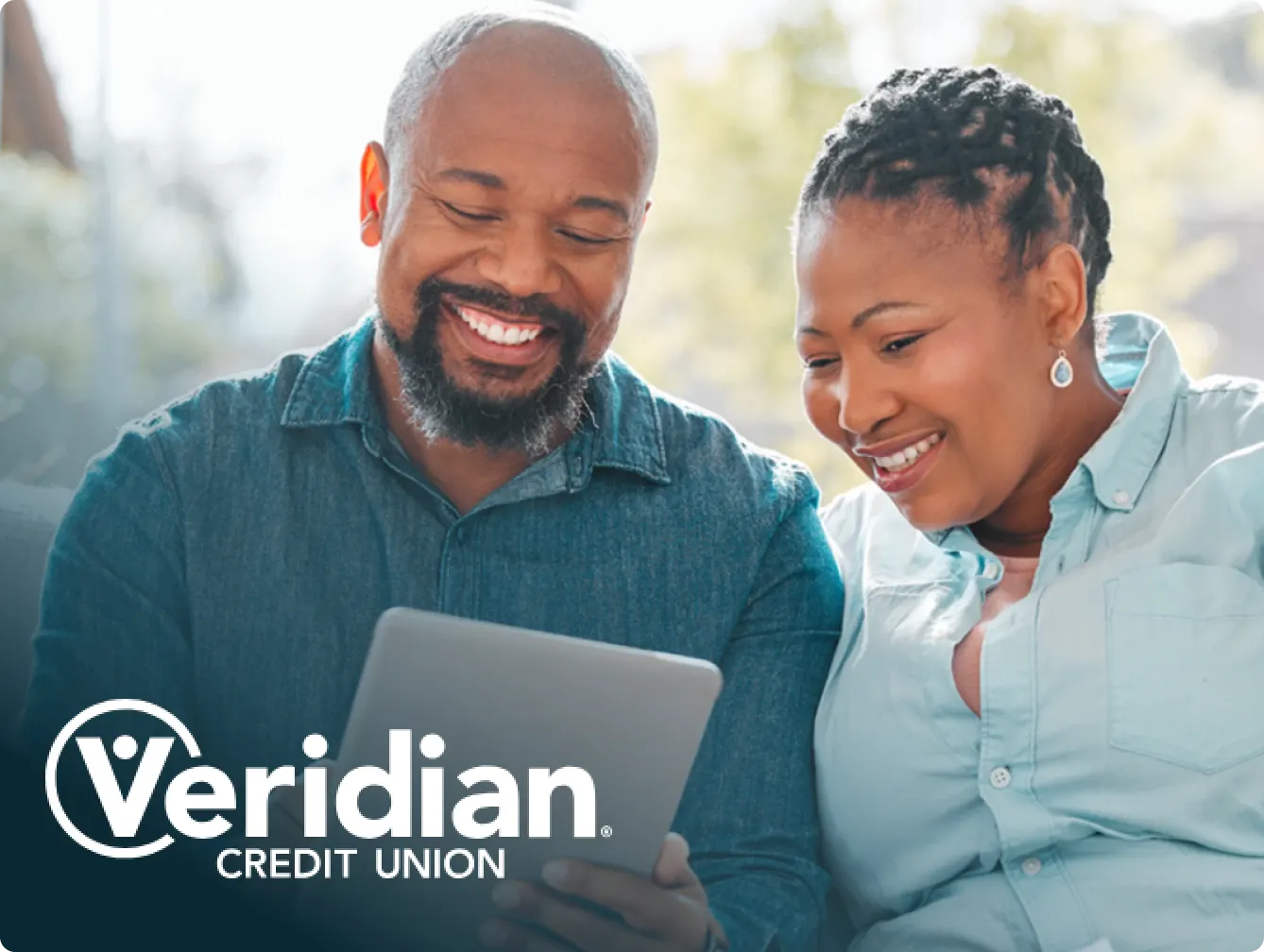 Veridian Credit Union advertisement featuring smiling people sitting together and looking at a tablet. The man is wearing a denim shirt, and the woman is wearing a light blue blouse. The Veridian Credit Union logo is prominently displayed at the bottom left of the image.