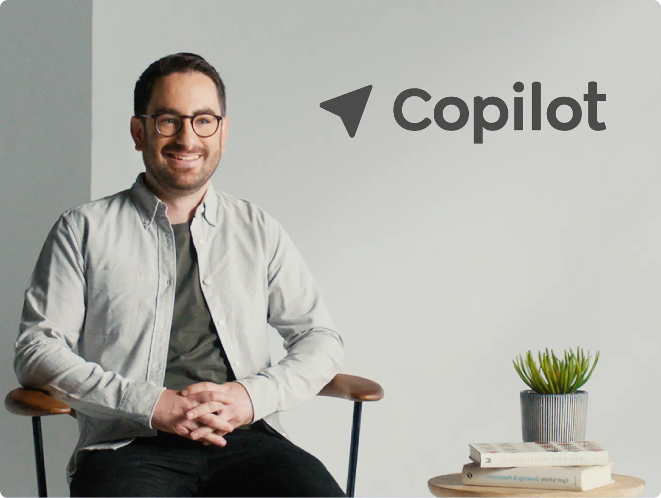 Copilot advertisement featuring a smiling man wearing glasses and a light gray shirt, sitting on a chair. Next to him is a small table with a potted plant and books. The Copilot logo is prominently displayed at the top right of the image on a light gray background.