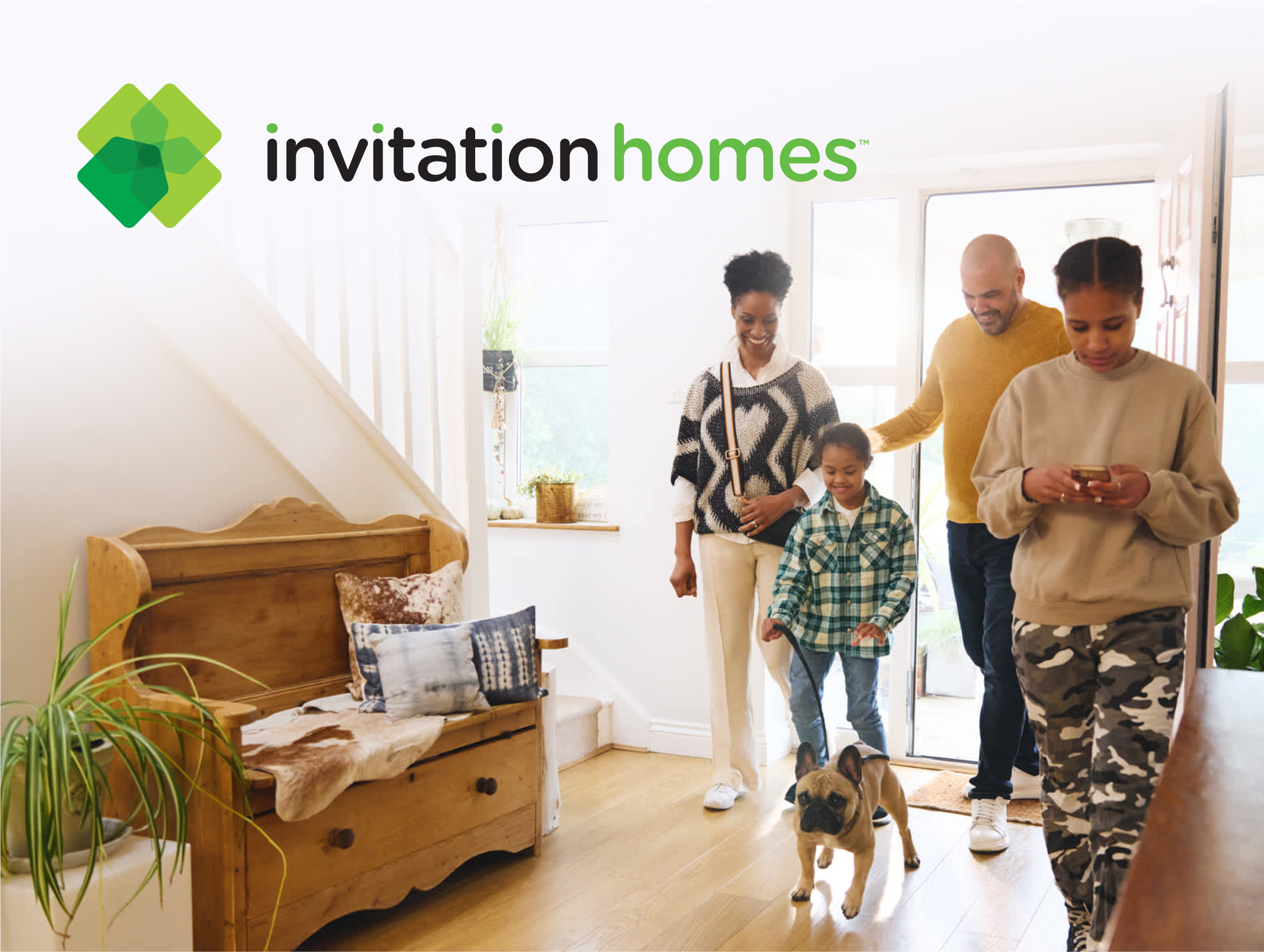 A family of four enters a bright, modern home with a small dog on a leash. The parents are smiling, while one child walks the dog and another checks their phone. The room features wooden flooring, a cozy wooden bench with decorative pillows and blankets, and green houseplants. The Invitation Homes logo is displayed prominently in the upper-left corner.