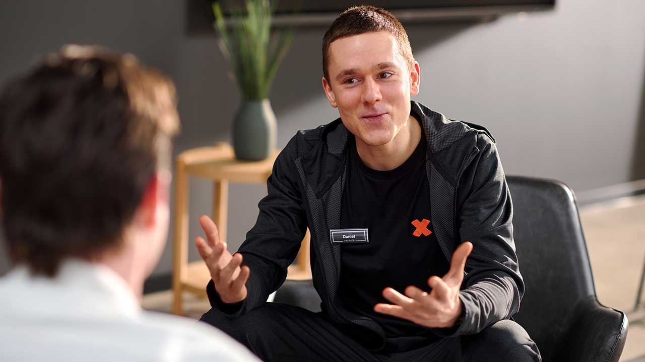 Bild:Ein FitX-Mitarbeiter spricht in einem Beratungsgespräch mit einer weiteren Person. Er lächelt und gestikuliert freundlich mit den Händen.