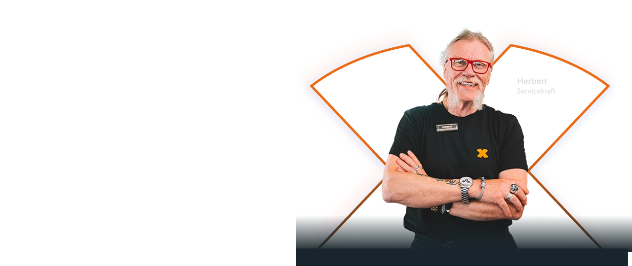 Bild: Eine männliche FitX Servicekraft steht frontal vor einem dunklen Hintergrund mit einer großen X‑Grafik und hat beide Arme vor dem Körper verschränkt. Die Person trägt ein schwarzes T‑Shirt mit FitX‑Logo und ein Namensschild.