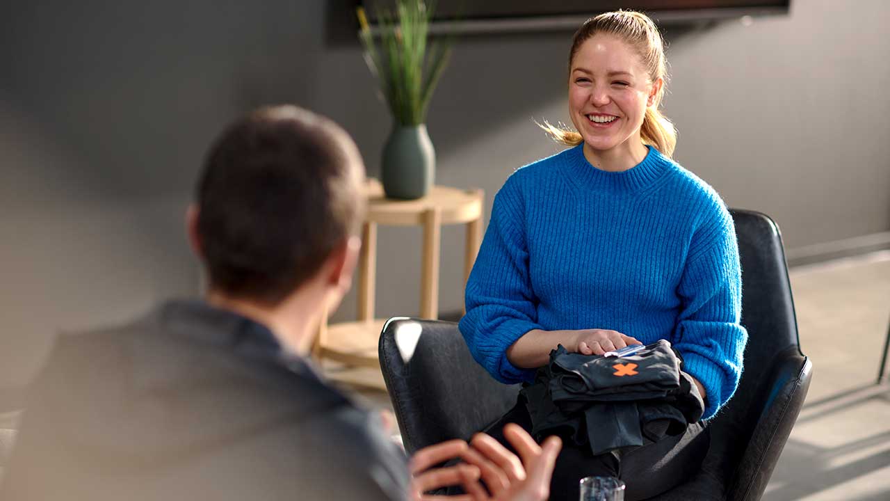 Bild:Eine junge Frau spricht mit einer weiteren Person. Sie trägt einen blauen Pullover, lacht herzlich und hält eine gefaltete schwarze Trainingsjacke mit orangenem FitX-Logo in den Händen.