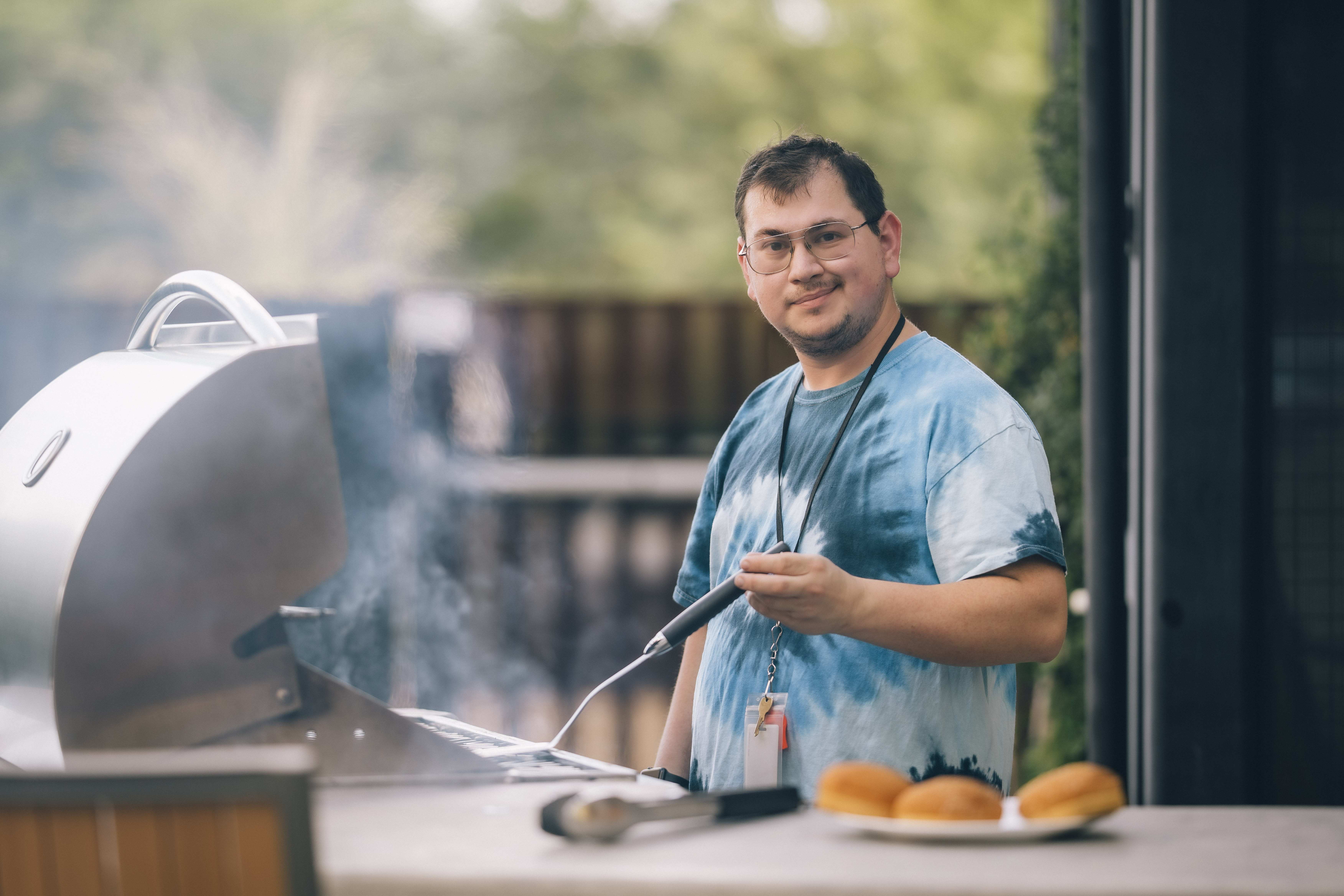 Eli holding a spatula standing in front of a smoking grill next to a plate of hamburger buns