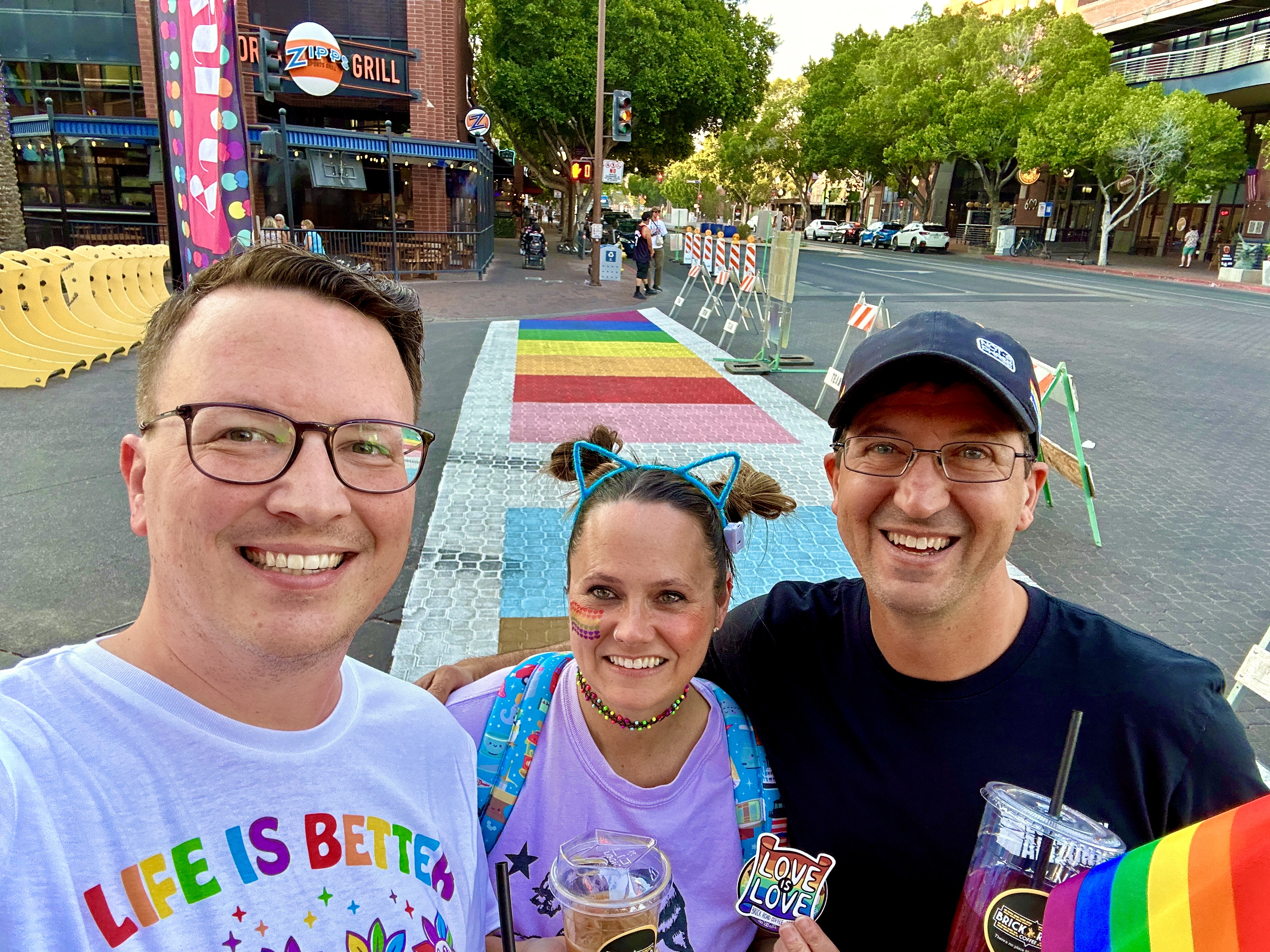 Three people in Pride outfits smiling for a selfie in front of a rainbow crosswalk
