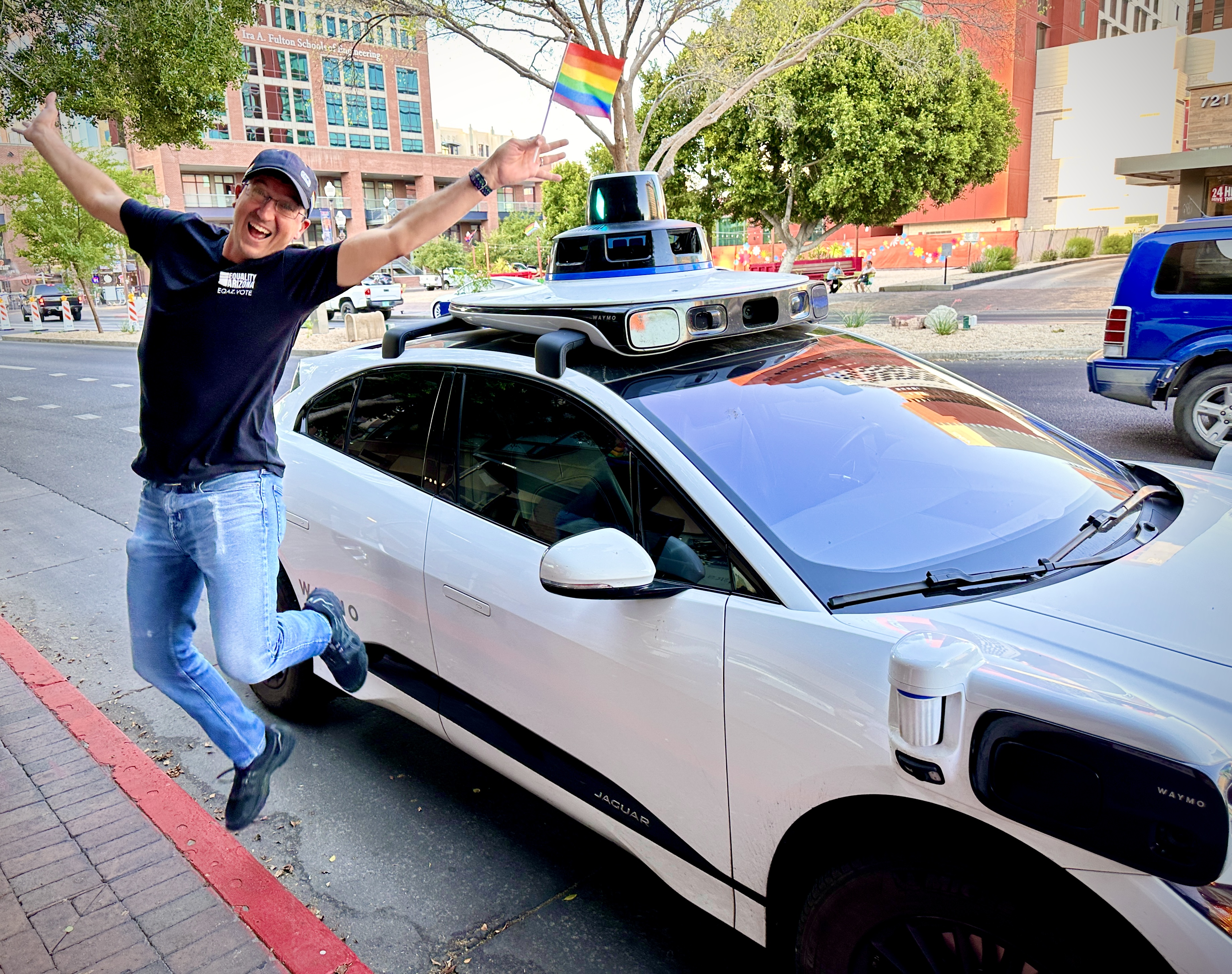 Man holding a rainbow Pride flag, jumping in the air in front of a parked Waymo vehicle