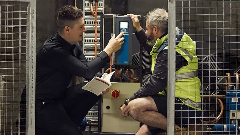 Two technicians inspecting electrical equipment in an industrial setting, one holding a tablet and the other in a safety vest.