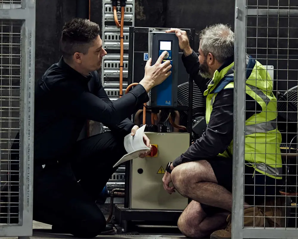 Two technicians working on industrial equipment with blue and black control panel in a facility with metal cages.