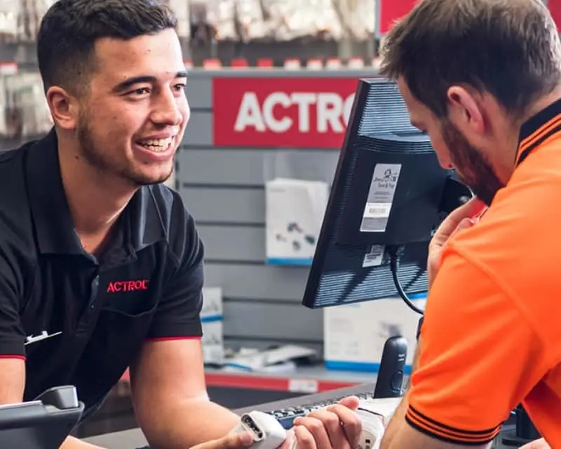 Employee in black polo shirt assisting customer in orange jacket at checkout counter with computer monitor