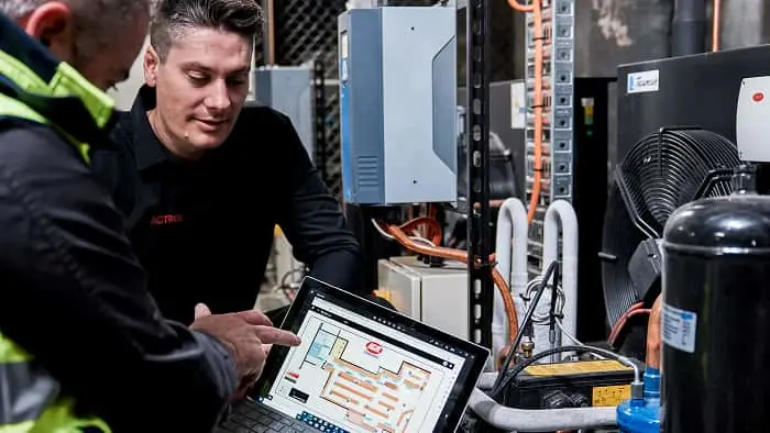 Two technicians examining a tablet displaying a technical diagram next to HVAC equipment with visible compressor and fan.