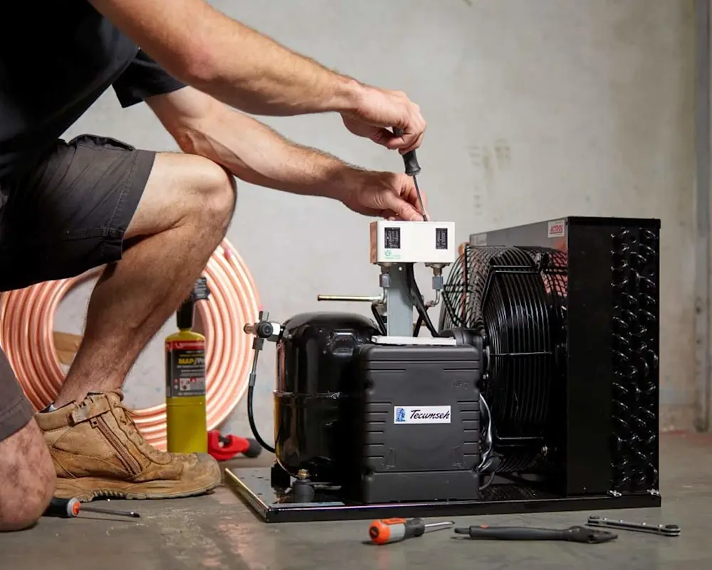 Person crouching while adjusting a black refrigeration compressor unit with tools on the floor nearby.