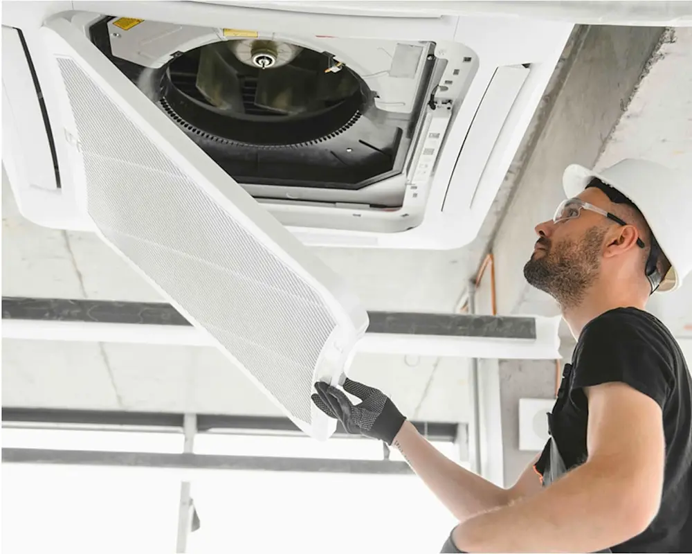 Technician wearing safety glasses inspecting an open ceiling-mounted air conditioning unit with exposed fan and filter.