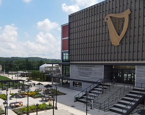 Exterior of Guinness Open Gate Brewery with a large golden harp logo on a dark modern building and outdoor seating area.