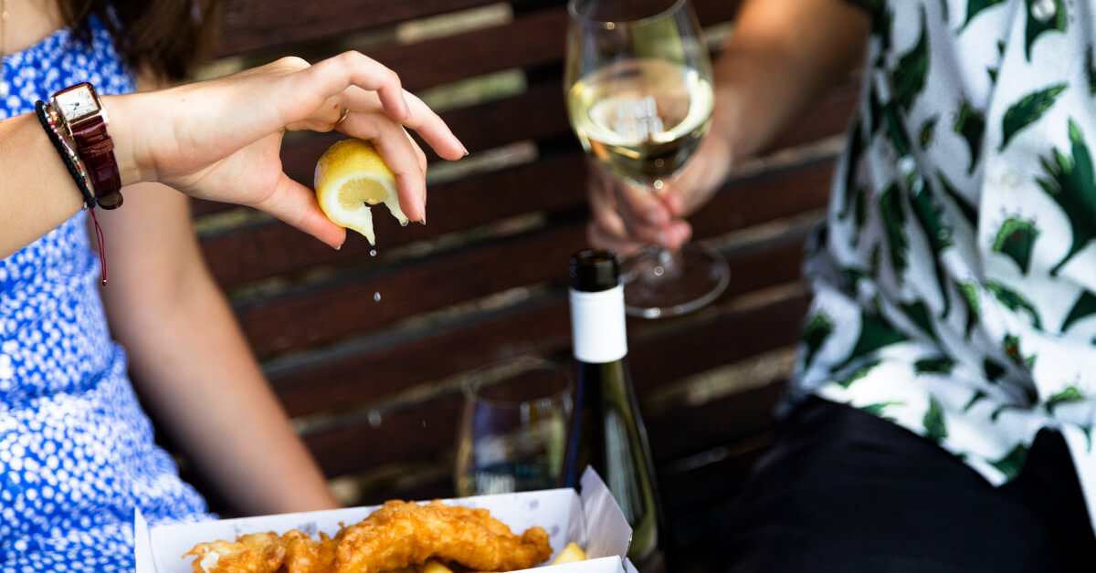 Image of someone squeezing a lemon wedge onto fish and chips, a bottle of riesling is in the background
